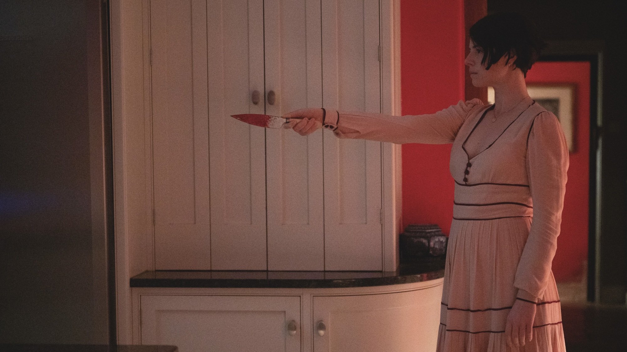 A woman in a pink dress points a bloody knife at an unknown intruder.