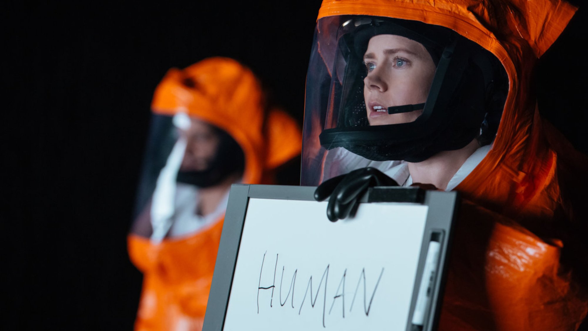 A woman in an orange hazmat suit holds up a whiteboard with "human" written on it