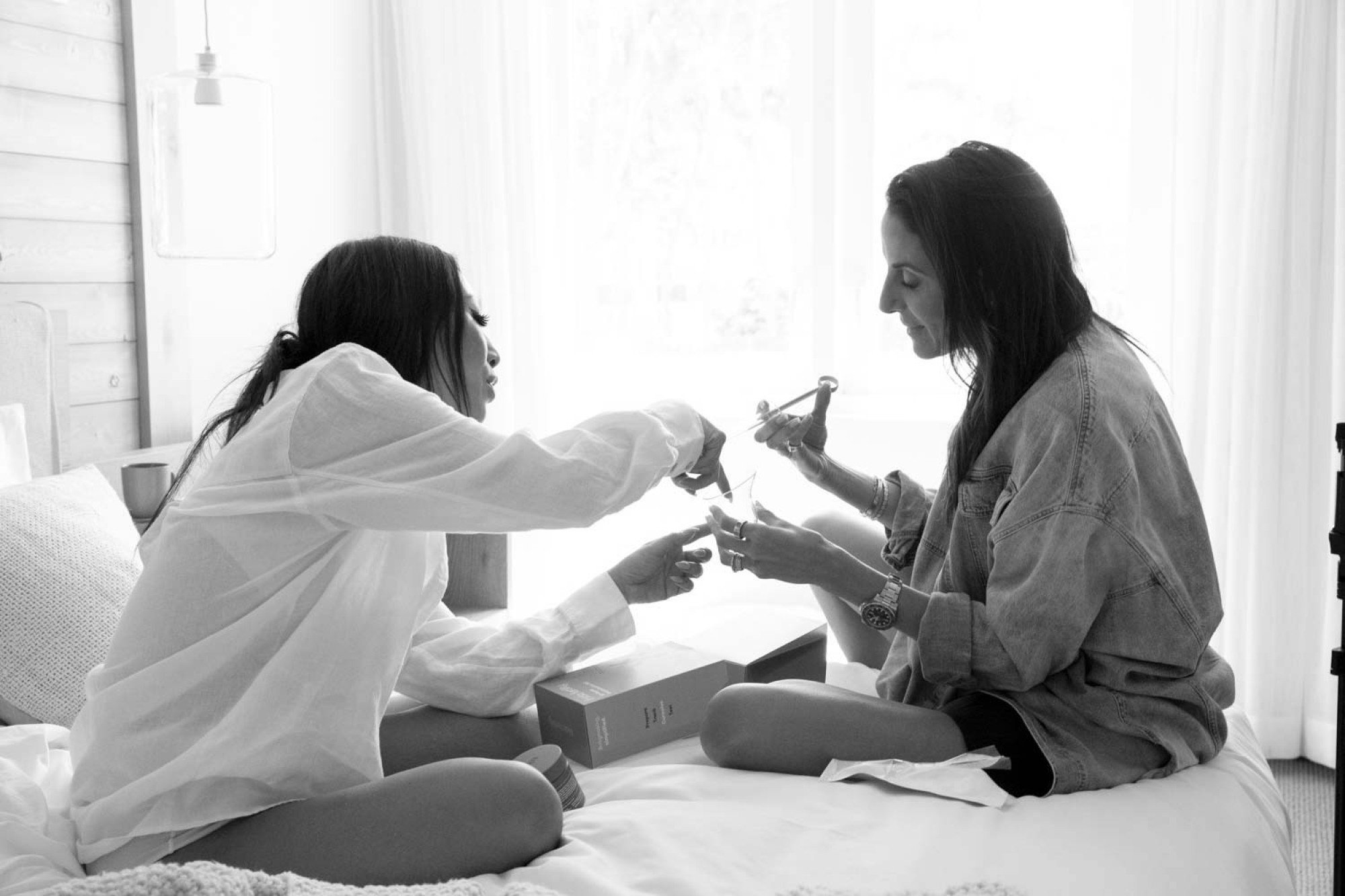 Two women sit in front of each other on a bed, holding and discussing Frida products.